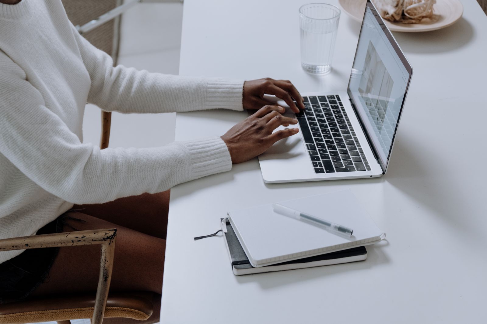 A person typing on a laptop beside a notebook at a table